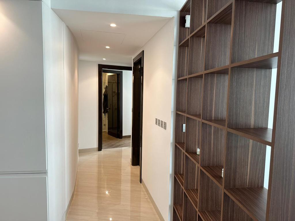 A well-lit hallway is depicted, featuring smooth marble flooring. To the right, a wooden shelving unit offers organized storage space, while doorways lead to various rooms at the far end. The walls are painted in neutral tones, contributing to a clean, modern aesthetic.