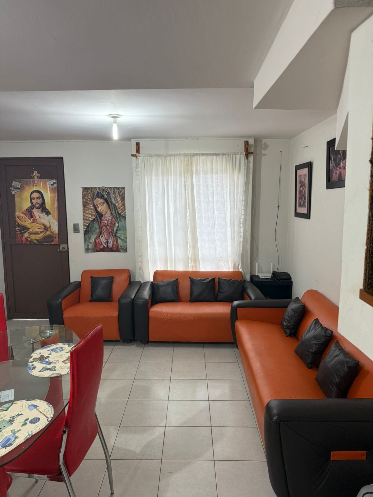 A living area showcases a combination of black and orange sofas arranged for comfortable seating. Artwork adorns the walls, contributing to the ambiance. Natural light filters through sheer curtains, illuminating the tiled floor and a nearby dining table set with red chairs.