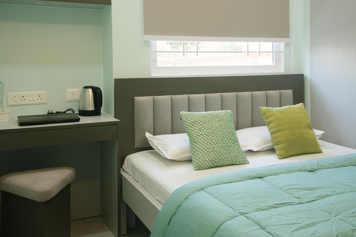 A serene sleeping area featuring a spacious bed with neatly arranged pillows and a lightweight quilt. A desk with a kettle is positioned nearby, complemented by a simple stool. Natural light enters through a window partially covered by a light blind.