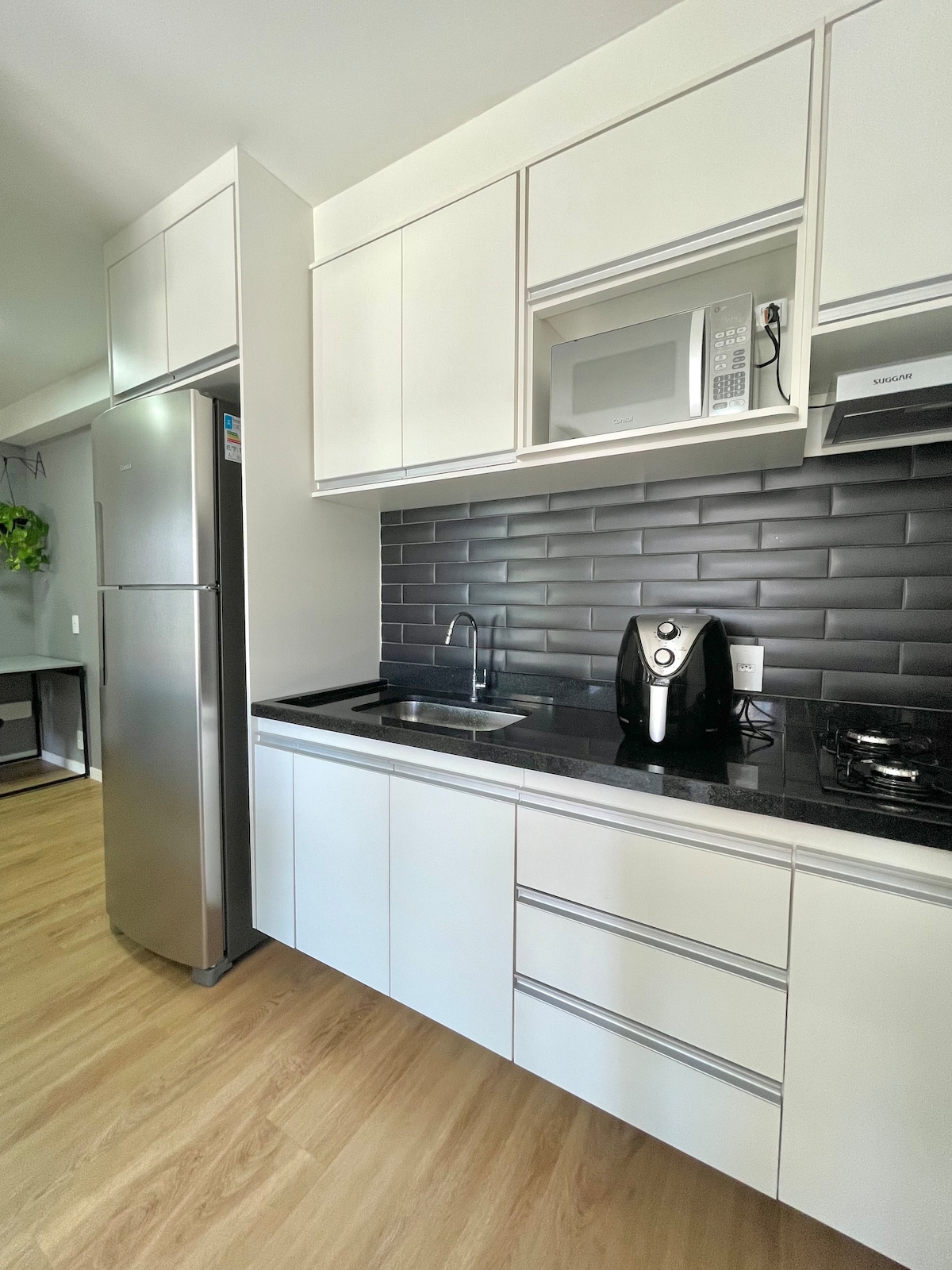 The kitchen features sleek white cabinetry complemented by a dark backsplash. A refrigerator, microwave, and coffee maker are visible, along with a sink and a stovetop. Wood flooring adds warmth to the modern space.