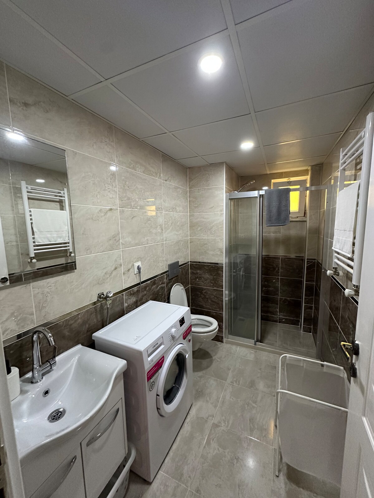 A modern bathroom features light-colored tiled walls and a spacious shower enclosure. A washing machine is positioned under the sink, alongside a vanity with a mirror above it. Soft overhead lighting provides illumination, while towels are neatly hung on a rack.