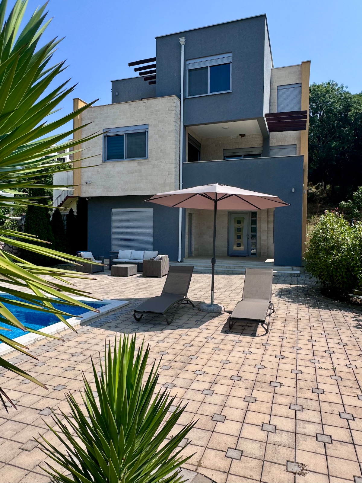 The modern villa is set against a backdrop of lush greenery, featuring a tiled outdoor area with lounge chairs under a large umbrella. A refreshing pool is visible alongside the patio, creating a relaxing environment for outdoor leisure.