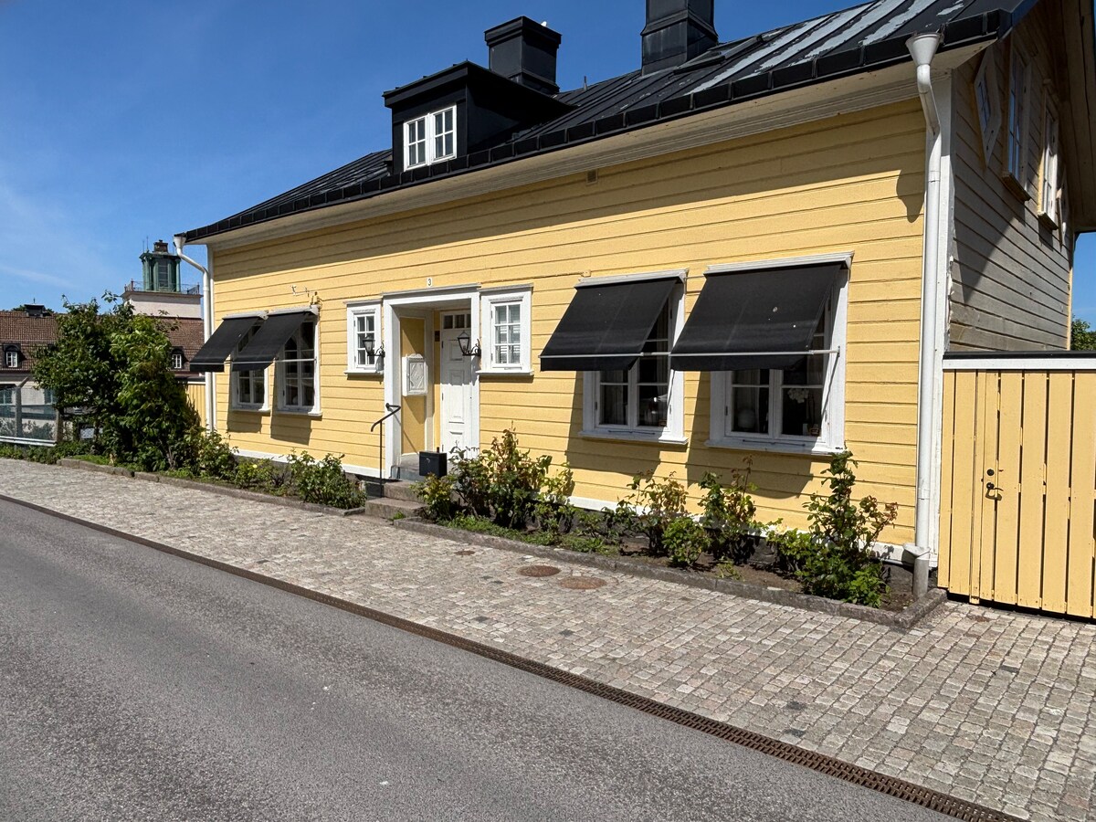 A bright yellow facade is seen, featuring multiple windows covered by window awnings. The entrance is adorned with steps leading to a white front door. Neatly trimmed bushes line the pathway, which is paved with stones and located along a quiet street.