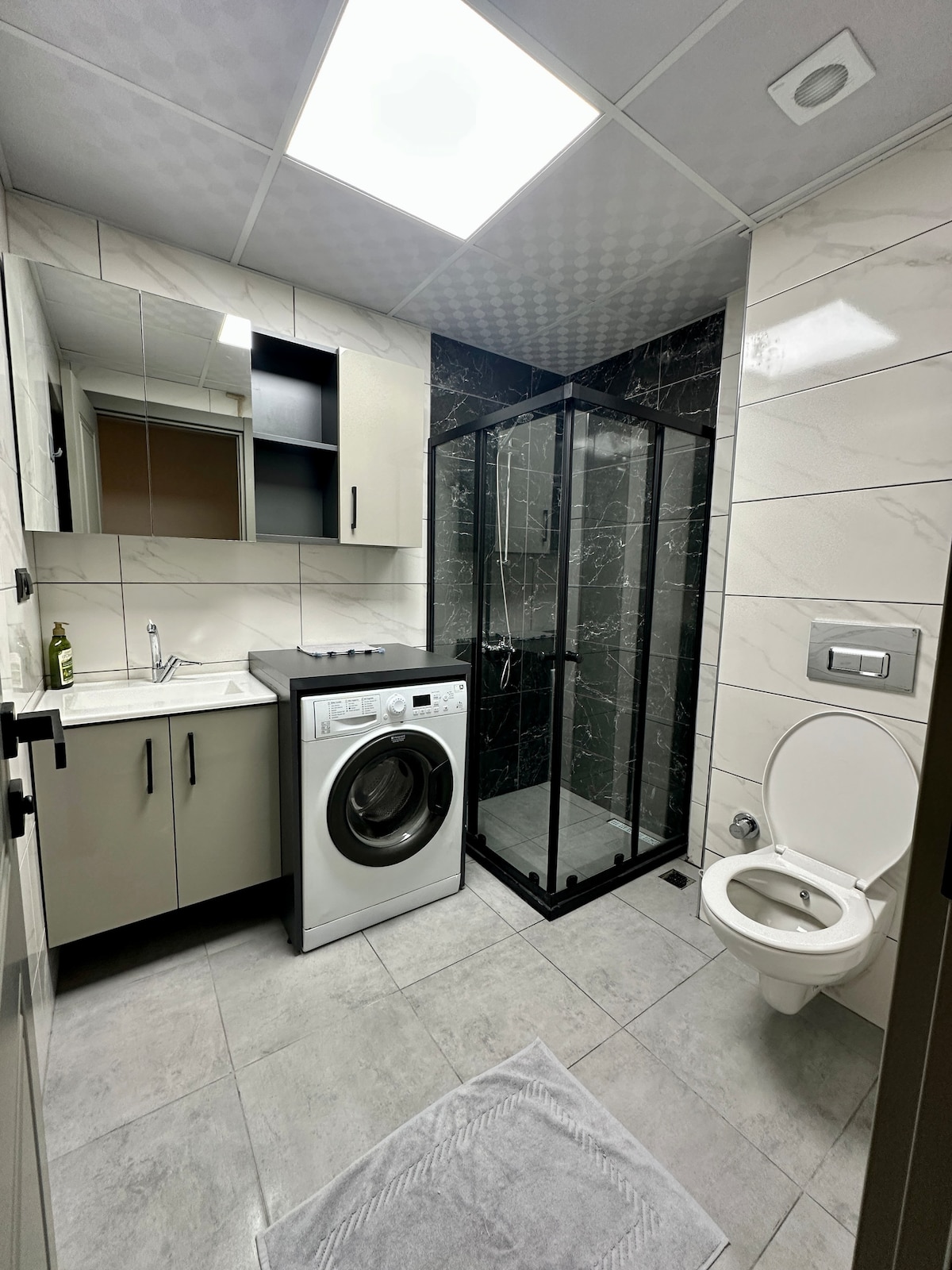 A modern bathroom features a sleek shower enclosure with black tile accents, a compact washing machine, and a white vanity with a mirror. The space is illuminated with overhead lighting, complemented by light grey tiles on the floor and a small white rug.