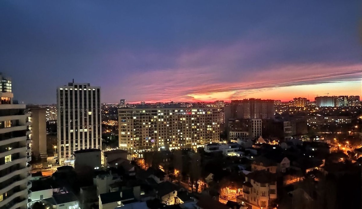 A city skyline is displayed at dusk, with a gradient of colors in the sky transitioning from orange to purple. Numerous buildings with illuminated windows are visible, creating a lively urban backdrop, while lower structures are partially shaded.
