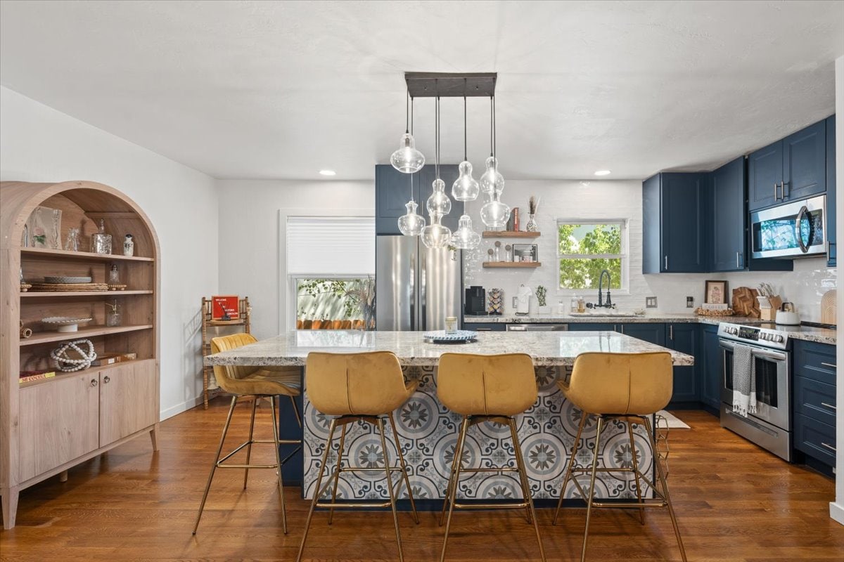 A modern kitchen showcases a large island with stylish bar stools, offering a comfortable space for gathering. Sleek blue cabinetry and stainless steel appliances enhance functionality. Distinctive tile patterns add visual interest to the countertop, while natural light filters through the nearby window, illuminating the space.