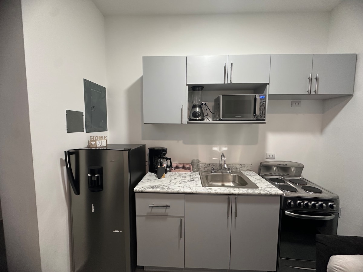 A compact kitchen area is displayed, featuring sleek grey cabinets and a countertop with a light marble finish. Appliances include a refrigerator, a coffee maker, a microwave, and a stove. Ample storage space is provided with cabinets and a sink.