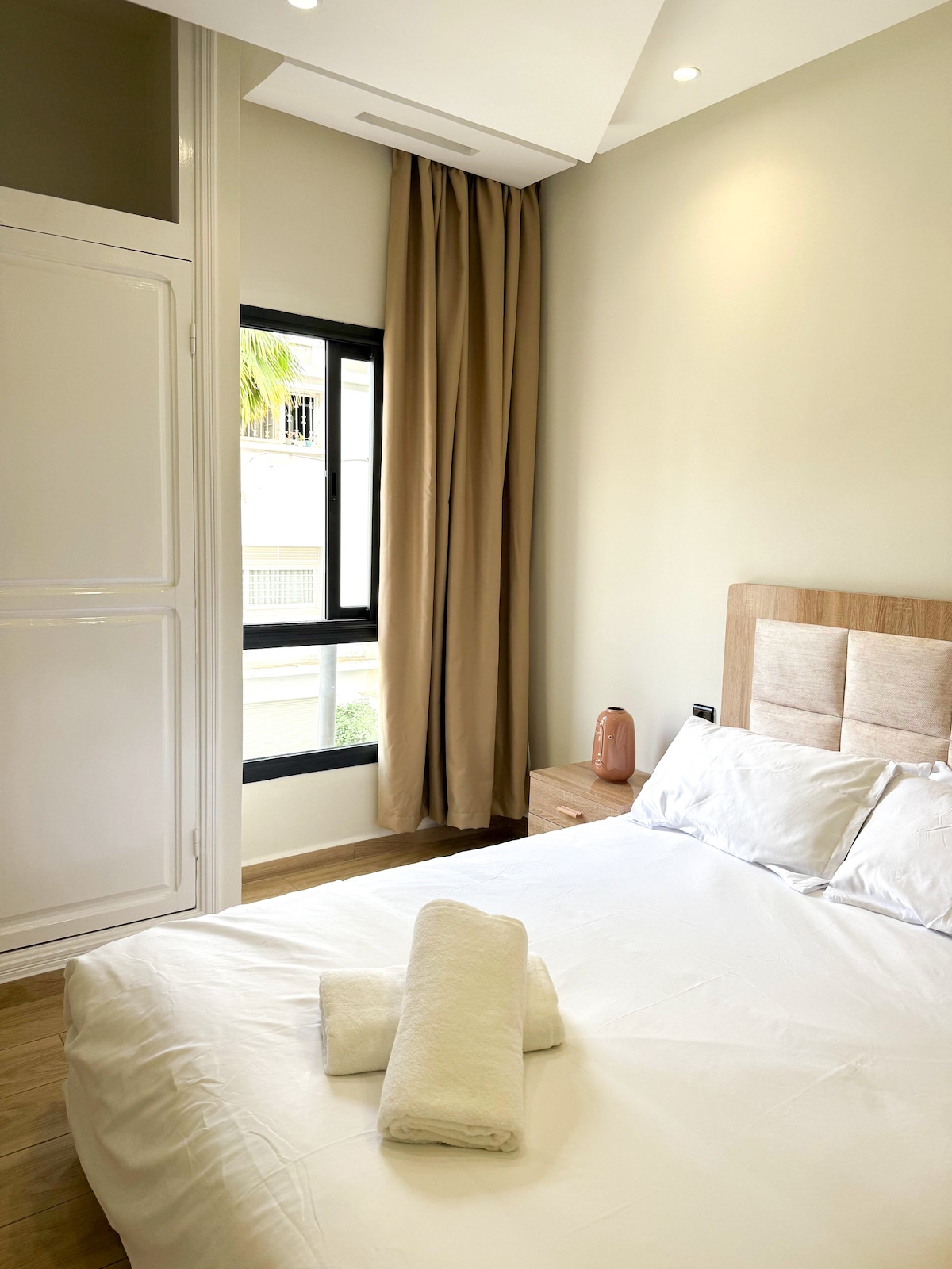 A modern sleeping area features a spacious bed with light bedding and two neatly arranged towels. A stylish headboard complements the decor, while natural light filters through a large window adorned with beige curtains.