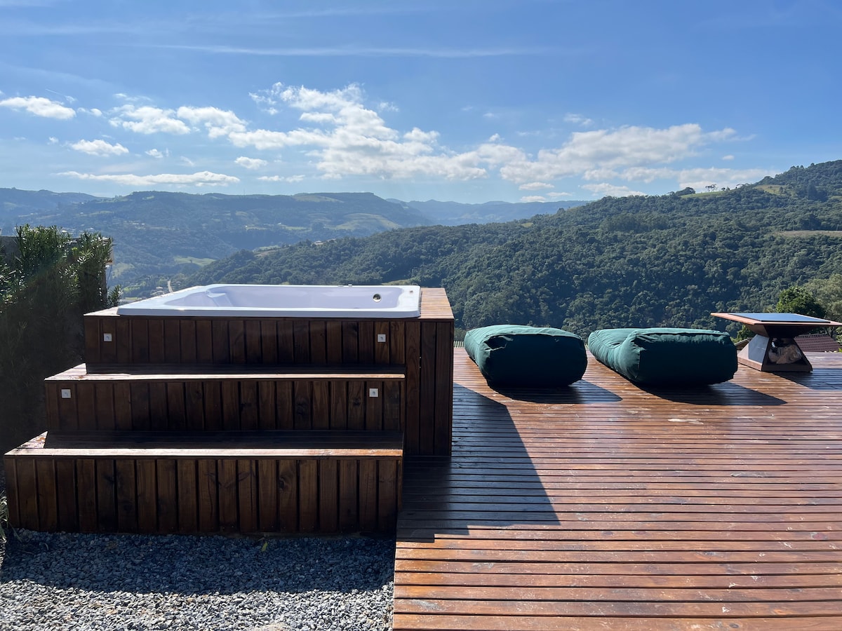An outdoor deck features a wooden hot tub positioned on tiered wood steps, overlooking a lush green landscape. Two plush seating areas are included, with deep green cushions adding comfort. The expansive view of rolling hills is highlighted against a clear blue sky.