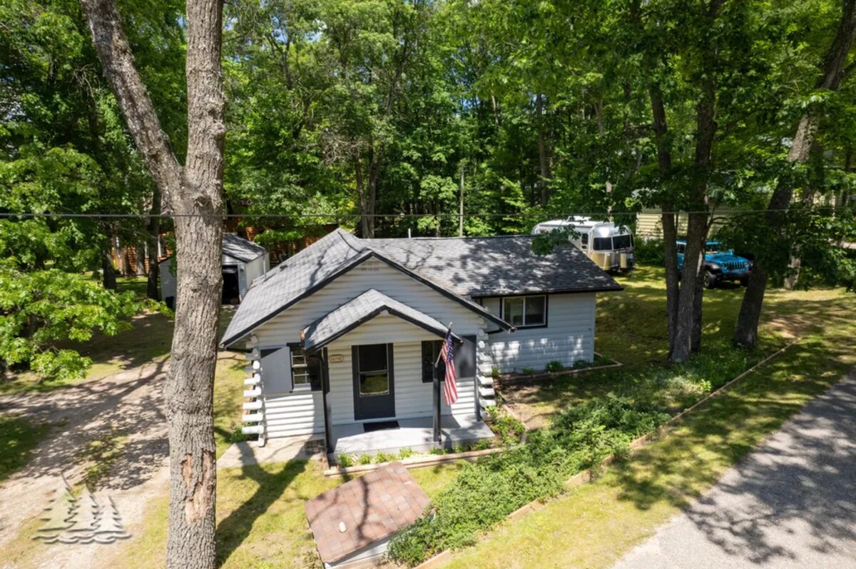The exterior of a cozy cabin is shown, surrounded by lush trees. A welcoming front porch features a small set of steps and a flag displayed. Gravel paths lead to the cabin, with additional outdoor space visible, creating an inviting atmosphere in a wooded setting.