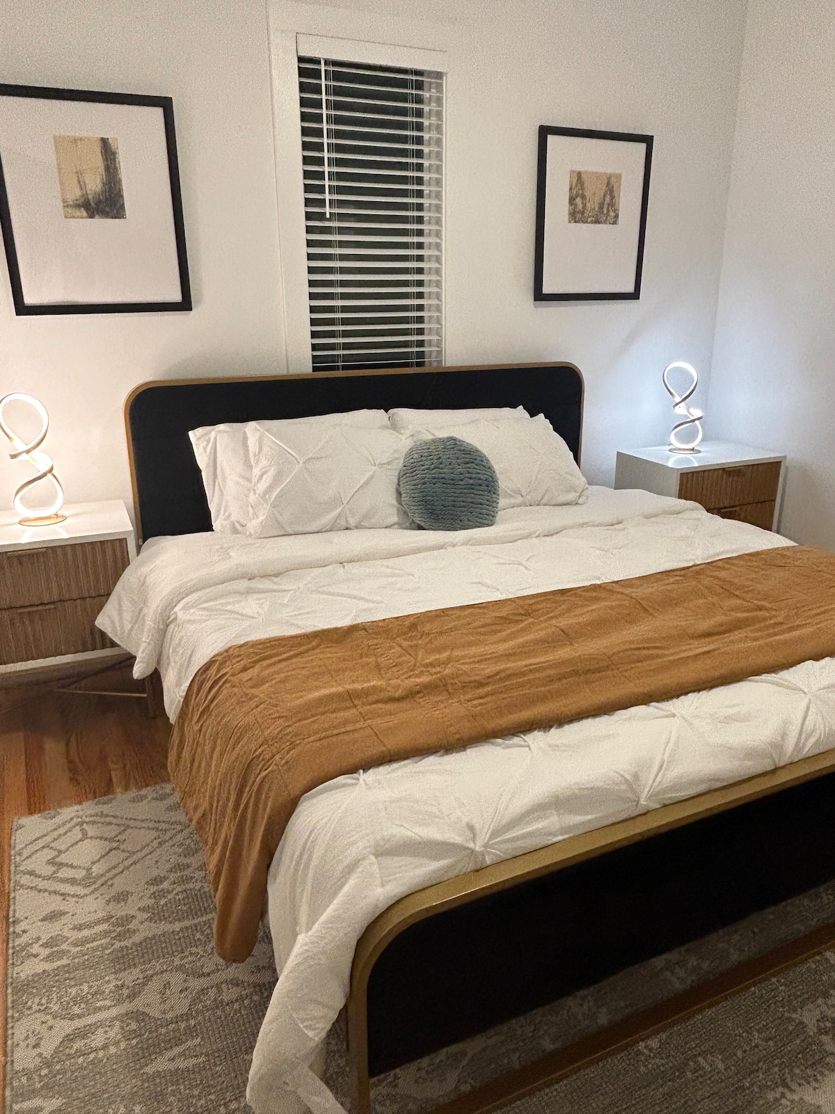 A well-lit bedroom features a spacious bed dressed in white linens, complemented by a warm-toned blanket. Two nightstands with modern lamps sit on either side of the bed. Artwork hangs on the walls, and natural light filters through a window with horizontal blinds.