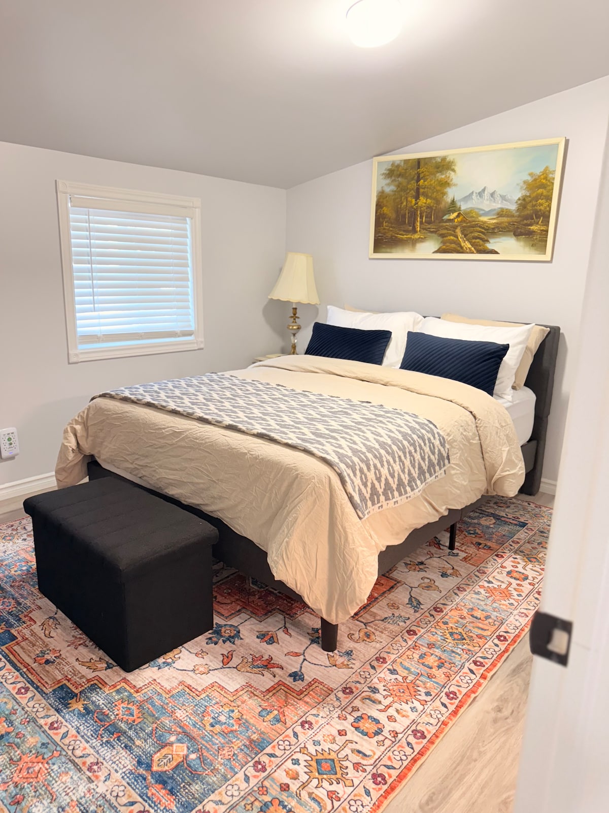 A cozy bedroom features a queen-sized bed dressed in soft linens, flanked by two pillows. A small bench is positioned at the foot of the bed. Light-colored walls complement a colorful area rug, and a landscape painting hangs above the bed, while natural light filters through a window with blinds.