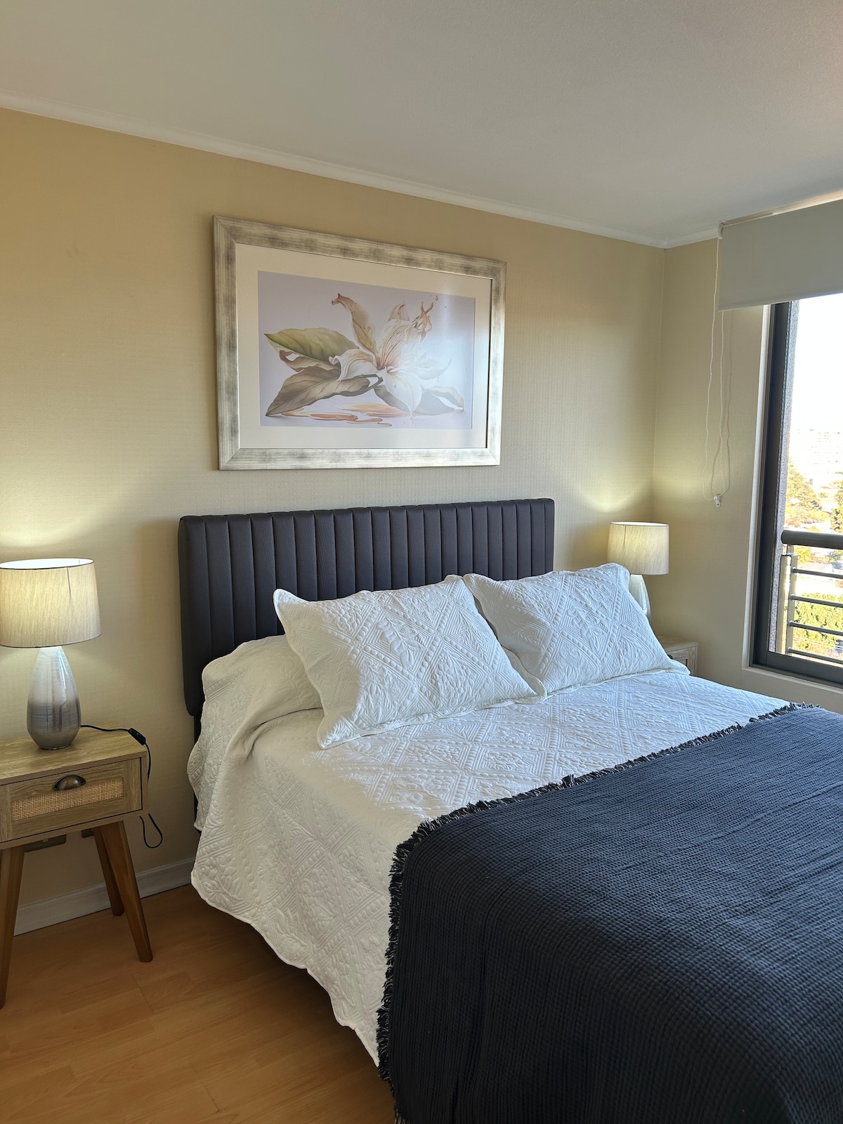A cozy bedroom is presented, featuring a spacious bed dressed in white linens and a dark throw blanket. A framed floral artwork adorns the wall above the bed. Two bedside lamps provide gentle lighting, complementing a small wooden nightstand beside the bed.