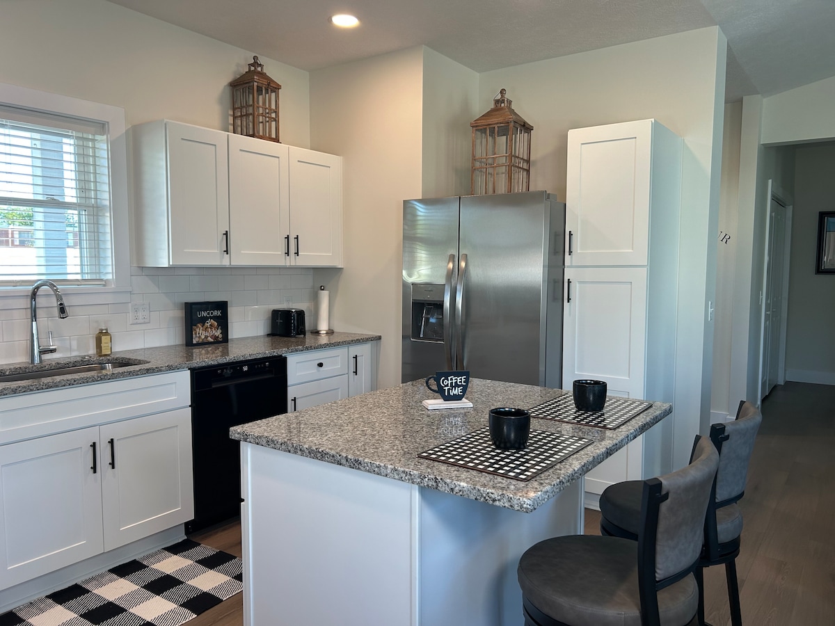 A modern kitchen showcases sleek white cabinetry and a spacious granite island. Stainless steel appliances, including a refrigerator and dishwasher, are visible. Two dark chairs are placed at the island, with a black and white striped placemat and mugs arranged on the counter.