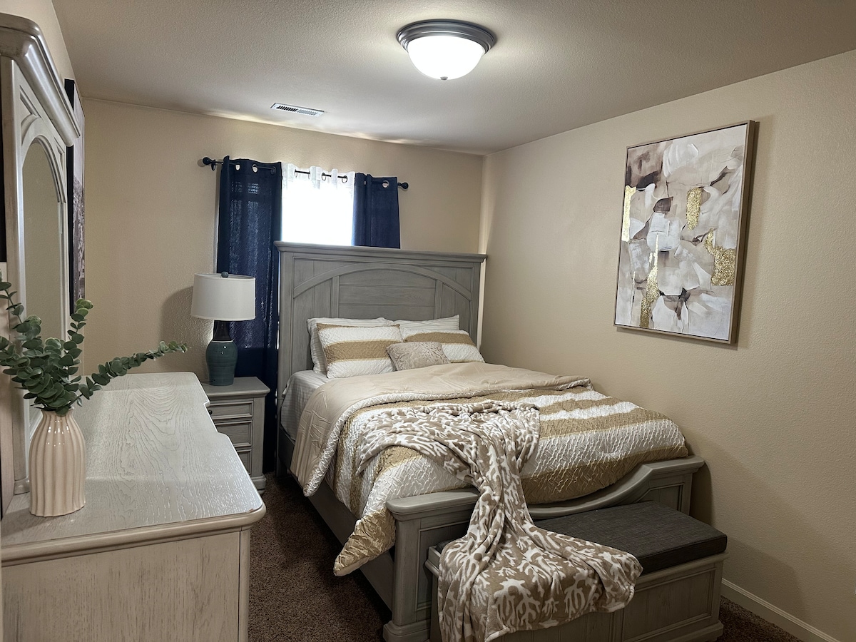 A well-appointed bedroom features a spacious bed with neutral-toned bedding, complemented by decorative throws. A bedside lamp and a small dresser are positioned beside the bed, while a wall art piece adds a touch of elegance. Natural light is filtered through sheer curtains.