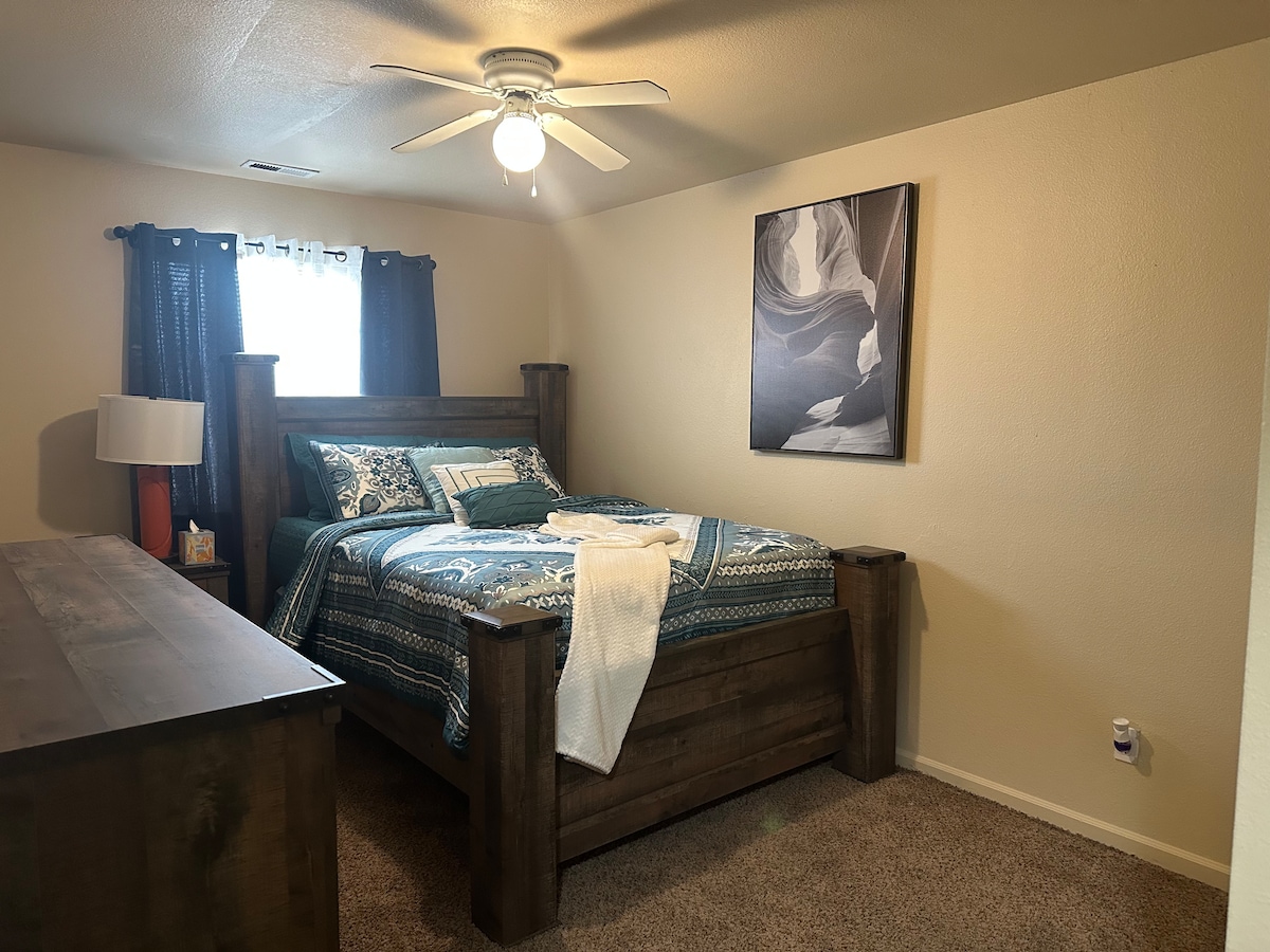 A cozy bedroom features a spacious wooden bed with a thick comforter. A bedside lamp provides light, while a piece of artwork adorns the wall above the bed. A dresser is visible beside the bed, enhancing the room's functionality.