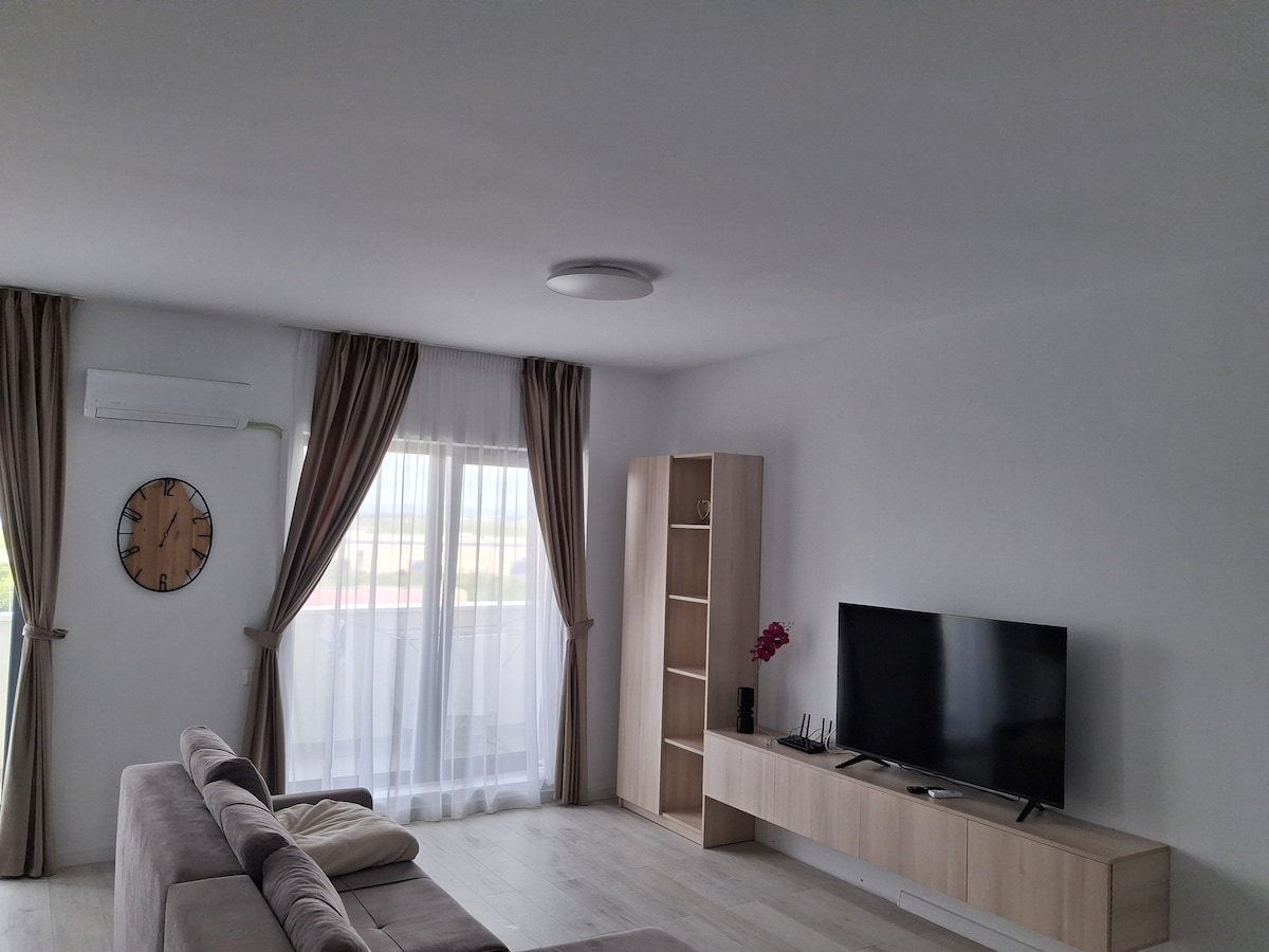A bright living area features a modern light-coloured bookcase and a television mounted on a low wooden cabinet. Floor-to-ceiling windows with sheer curtains allow natural light to fill the space. A comfortable sofa is positioned to create an inviting environment.