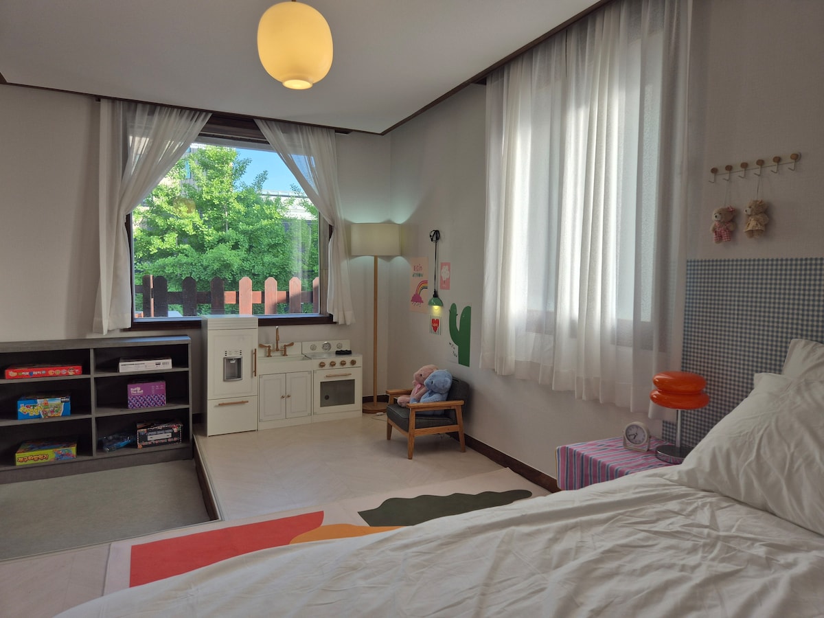 A spacious sleeping area features soft white curtains framing a large window, allowing natural light to fill the room. A play kitchen set sits among colorful toys on a low shelf. A cozy armchair is positioned nearby, offering a comfortable spot for relaxation.