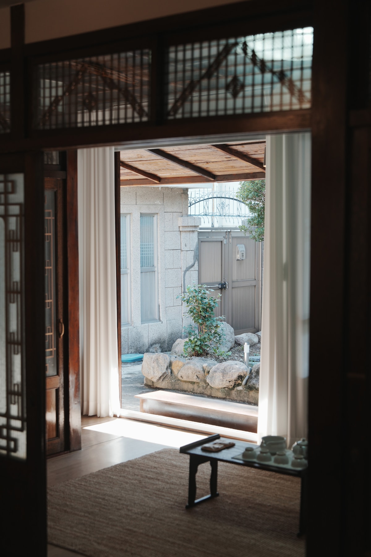 A view showcases an inviting entrance leading to a serene outdoor area. Natural light filters through sheer curtains, illuminating a stone bench surrounded by greenery. A traditional tea set is displayed on a low table, reflecting the tranquil atmosphere of the space.