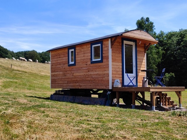 A wooden caravan is set on a raised deck, surrounded by a grassy landscape. Two blue chairs and a small table are positioned on the porch, inviting relaxation outdoors. A gentle hillside is visible in the background, with grazing sheep adding to the serene atmosphere.