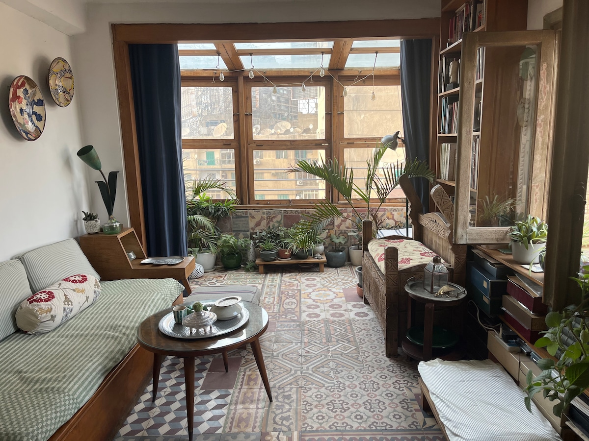 A welcoming living area features an L-shaped cotton sofa and a wooden bench adorned with plants. Decorative items include vintage plates on the wall and a bookcase filled with books. Natural light filters through large windows, casting a warm glow on the patterned rug.