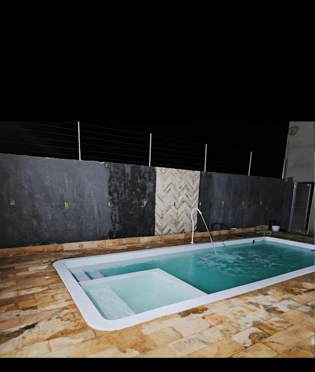A private pool is visible in a serene outdoor space, featuring clear water and a gently sloping shallow area. Surrounding walls are dark and textured, with a contrasting light-colored pattern on one side. The area is paved with natural stone tiles.