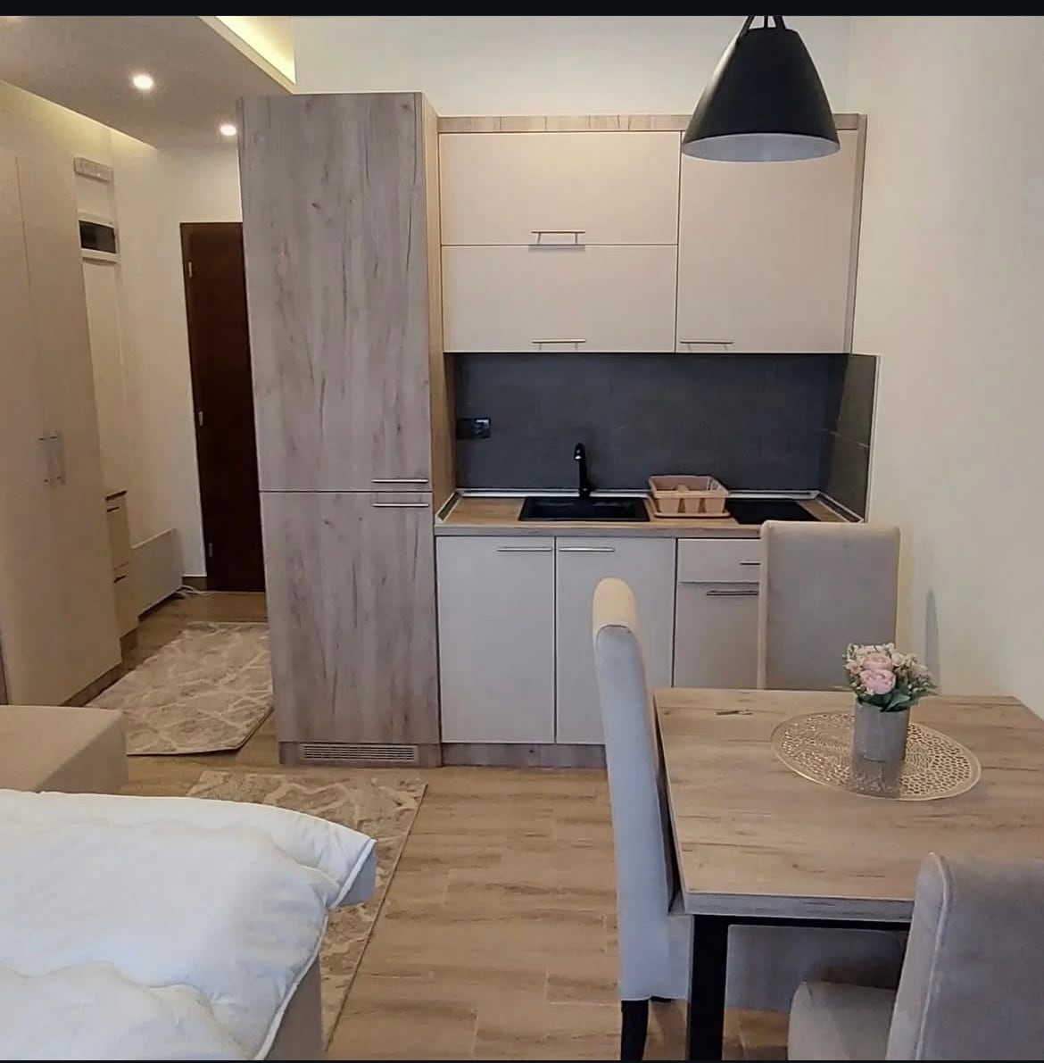 A modern kitchenette is featured with light cabinetry and a dark countertop, showcasing a sink and minimal appliances. A small dining table with chairs is positioned nearby, complemented by a decorative vase holding fresh flowers, contributing to the functional layout.