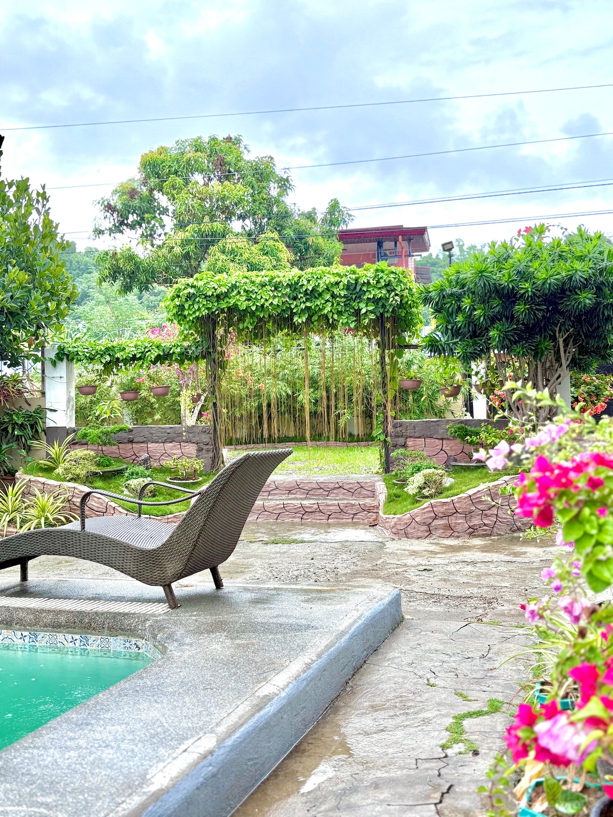 A serene garden area is depicted, featuring a path bordered by vibrant greenery and blooming flowers. A lounge chair provides a spot for relaxation near a tranquil pool, while a bamboo structure creates a natural archway leading into the lush surroundings.