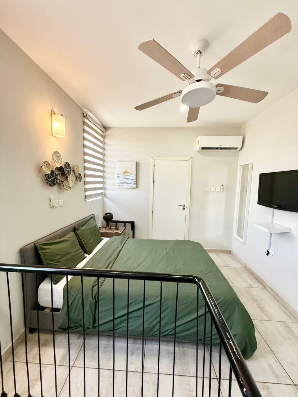 A modern bedroom features a spacious bed dressed in a dark green duvet. Wall art is displayed alongside a mounted television. Natural light enters through side windows with sleek blinds, while a ceiling fan and air conditioning unit are present for climate control.