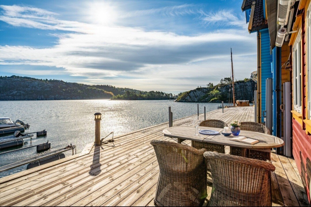 A wooden deck extends over the water, featuring a round table surrounded by wicker chairs. Sunlight reflects off the calm surface of the water, while the surrounding landscape includes gentle hills and a few boats moored along the dock.