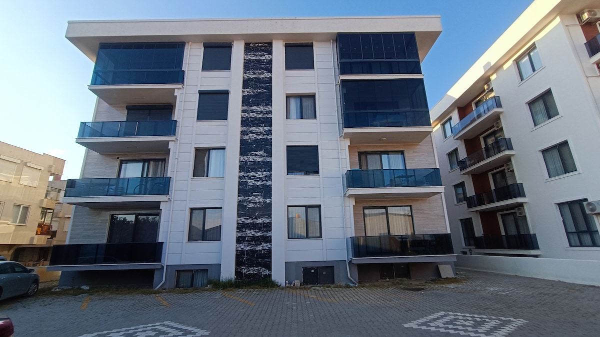 A modern three-story building is depicted, featuring a striking blue and white facade. Large glass balconies are seen on each level, enhancing the aesthetic appeal. The surrounding area displays a paved parking lot with clear spaces, contributing to the overall neat appearance of the property.