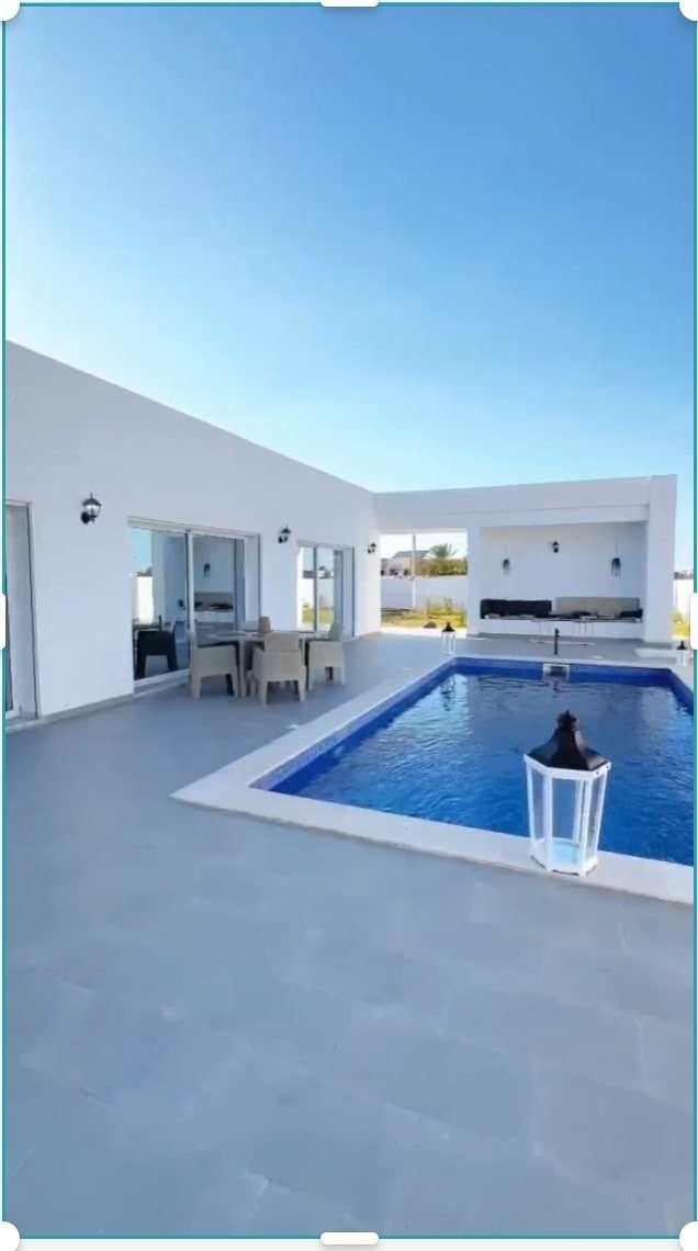 An outdoor area features a clear blue swimming pool bordered by a smooth patio. Nearby, a seating area is visible with several chairs surrounding a table. Large glass doors lead to an indoor space, allowing natural light to fill the area.