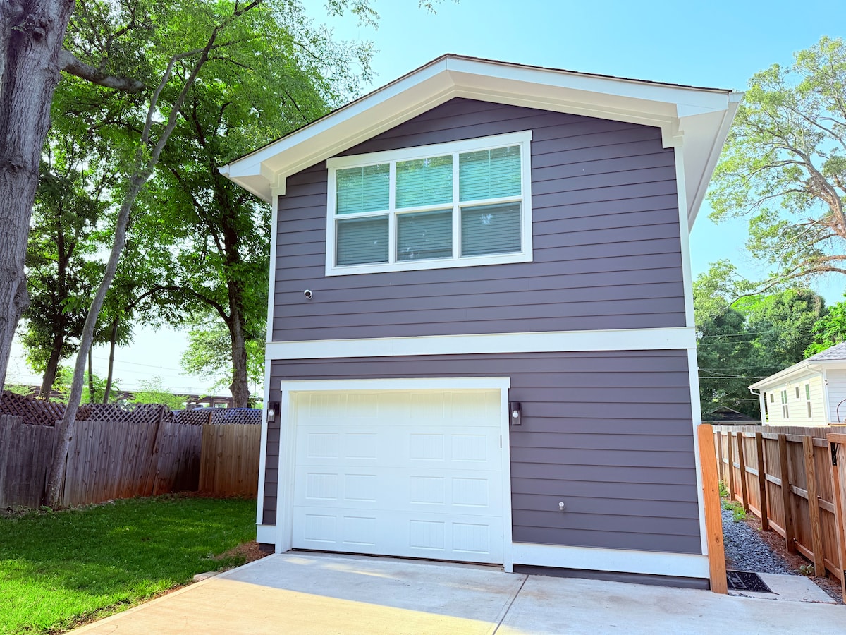 A modern two-story studio apartment is displayed, featuring a light gray exterior with white trim. Large windows allow natural light to flood in. A spacious garage with a white door is visible below. A well-maintained grassy area and a wooden fence surround the property.