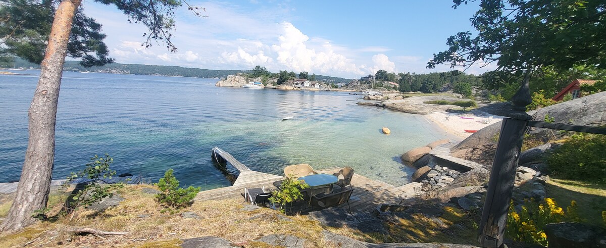 A serene waterfront view captures a sloping shoreline with clear, calm waters. A small dock extends into the water, bordered by smooth rocks. Lush greenery and scattered boulders frame the scene, while small boats are anchored nearby against the backdrop of distant hills and a partly cloudy sky.
