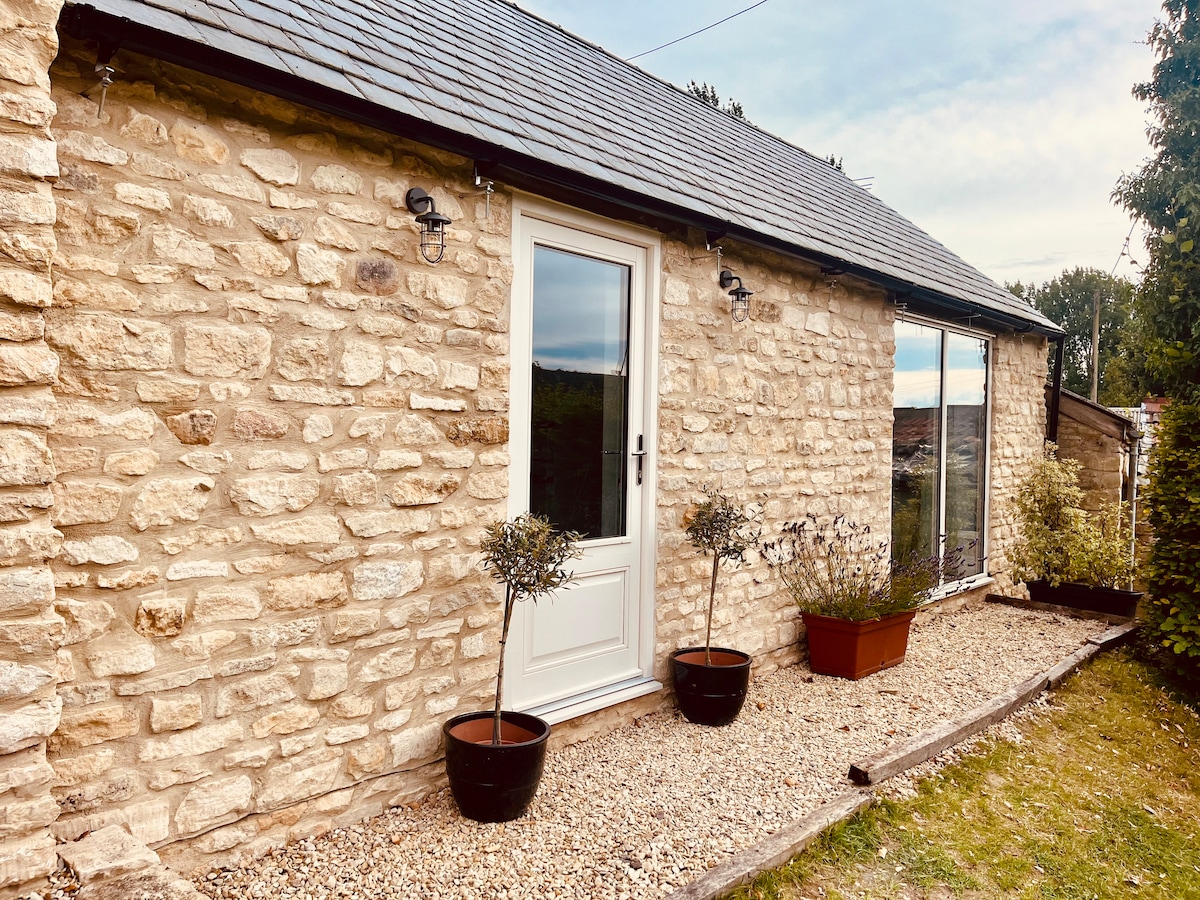 The exterior of a stone-built annexe is displayed, featuring a light-colored door framed by large windows. Planters with small trees are positioned on either side of the entrance, while gravel lines the pathway, complementing the rustic stone walls and sloped roof.