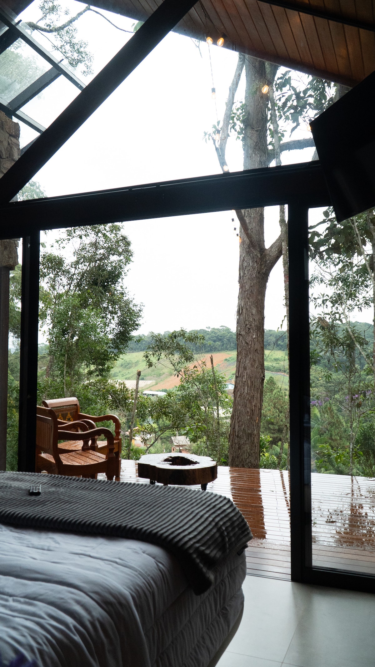 A spacious sleeping area features large glass doors that open to a deck surrounded by lush trees. A wooden chair is positioned on the deck, providing a view of the natural landscape. Soft bedding and a throw blanket are elegantly arranged on the bed.