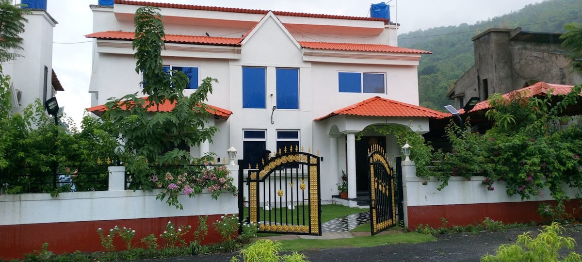 A two-story villa features distinctive architectural details, with a red-tiled roof complementing white walls. A wrought-iron gate stands at the entrance, surrounded by lush greenery and flowering plants, enhancing the inviting atmosphere of the property located at the mountain's base.