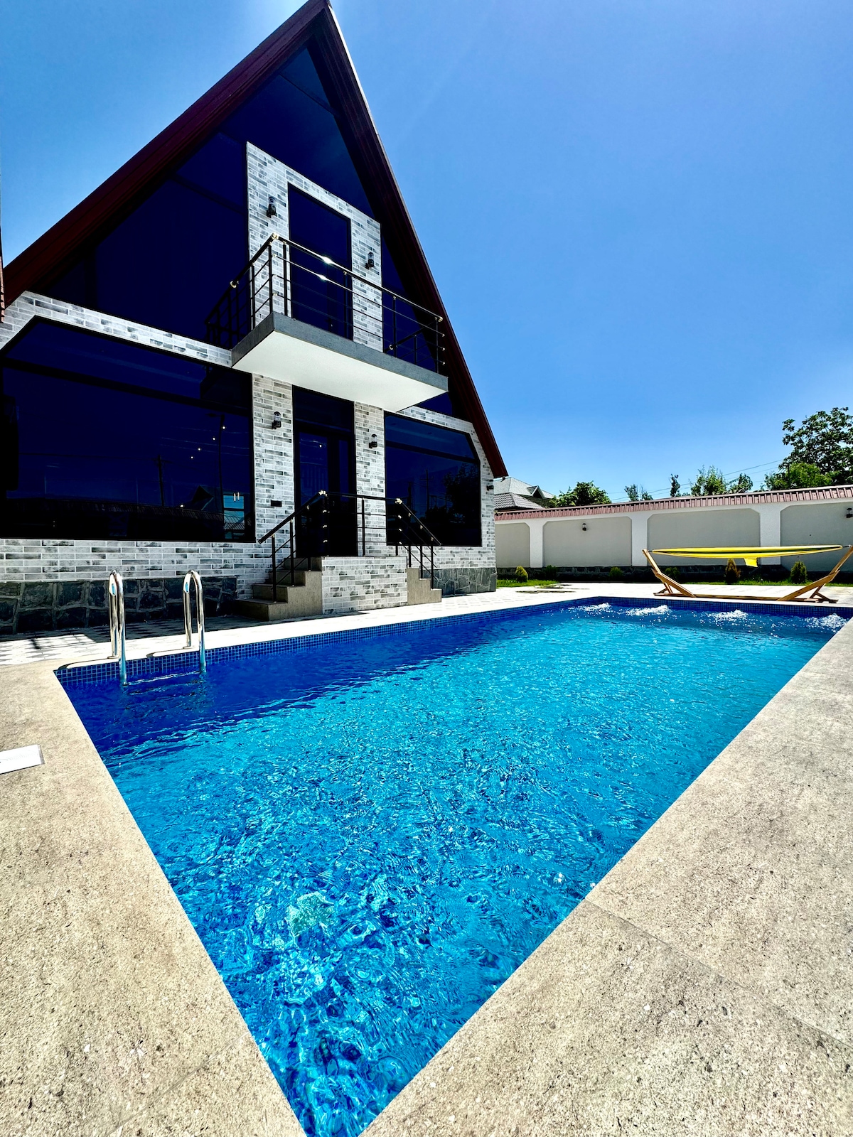A modern house is framed by large glass panels that reflect the clear sky. A serene pool is situated in the foreground, showcasing sparkling blue water and a textured stone deck. Two lounge chairs are positioned beside the pool, inviting relaxation.