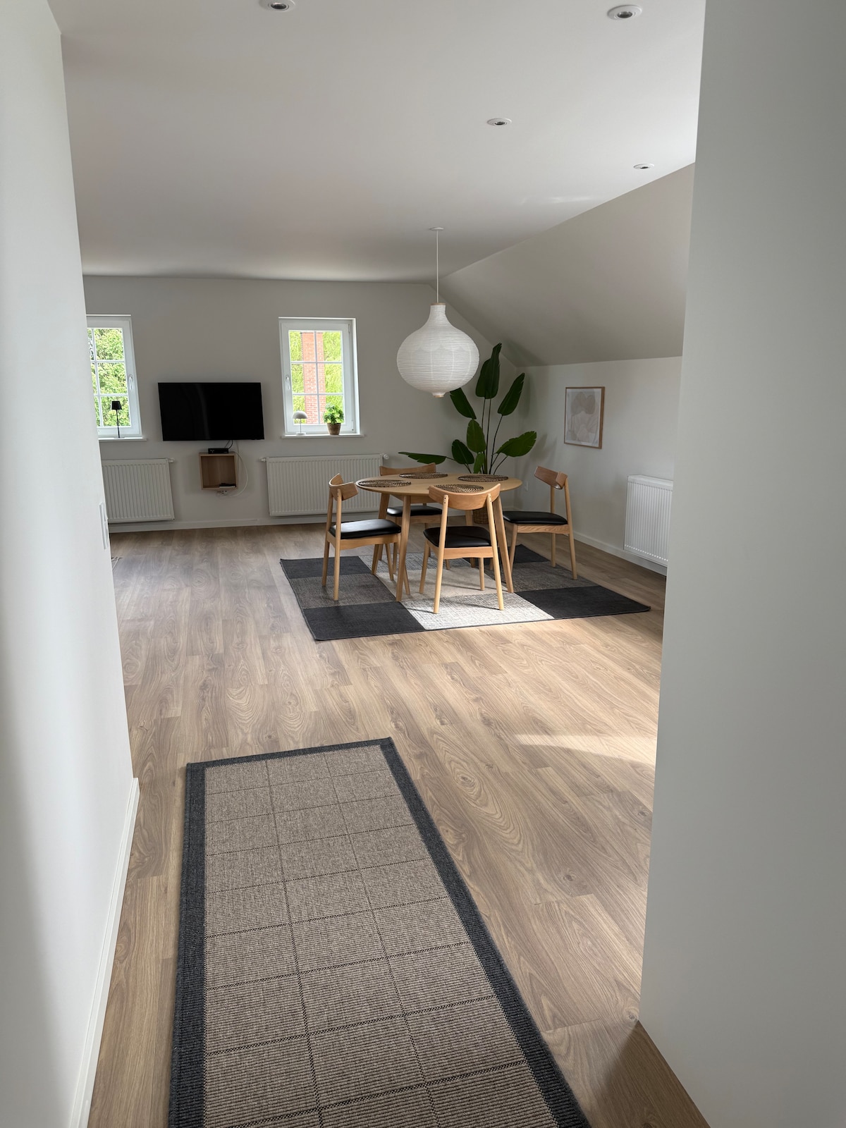 A well-lit area is viewed from a hallway, featuring a dining table surrounded by four chairs on a light-colored floor. A flat-screen television is positioned on the wall, and two windows allow natural light to fill the space. A plant in the corner adds a touch of greenery.