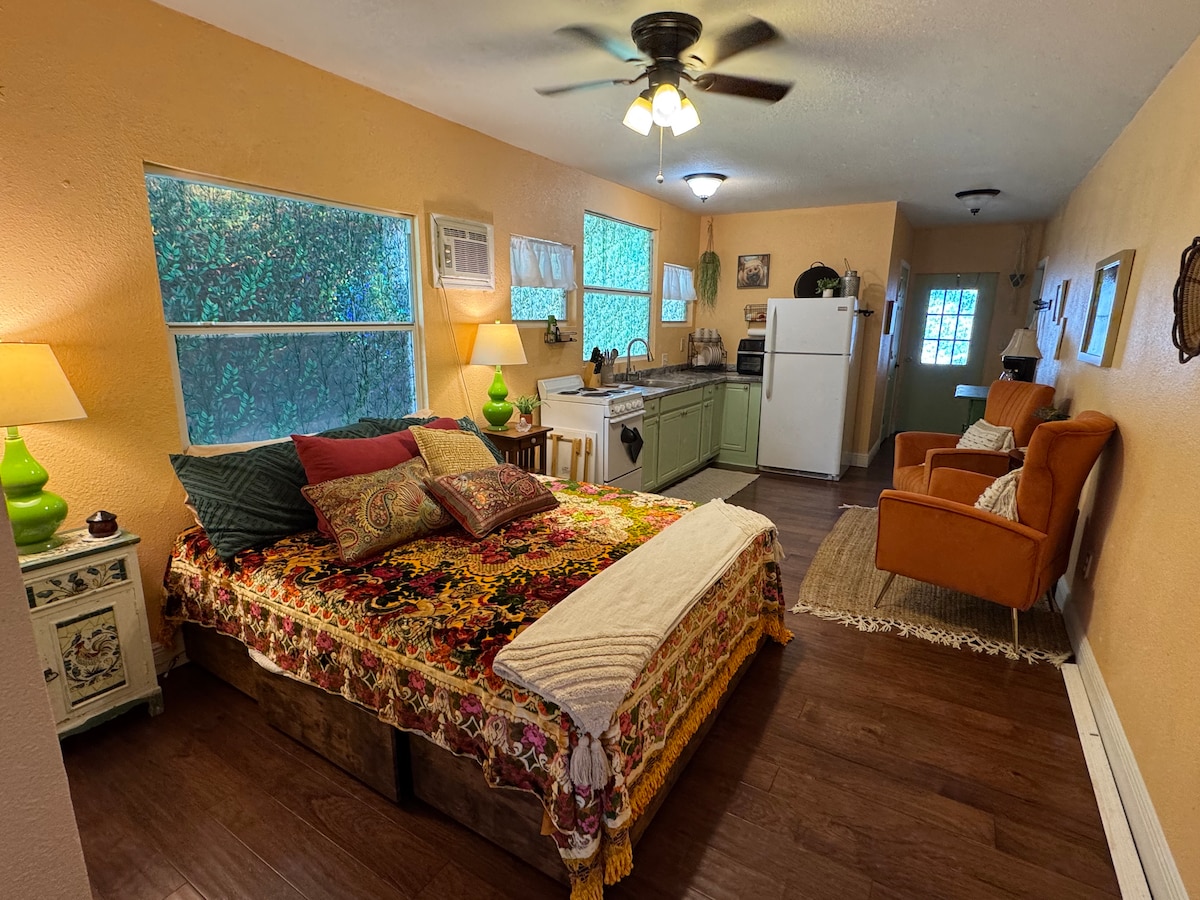 A cozy interior features a spacious bed adorned with a vibrant patterned blanket, complemented by accent pillows. The kitchen area includes a refrigerator and stove, with soft light coming through large windows. Warm orange walls and decorative touches create a unique atmosphere.