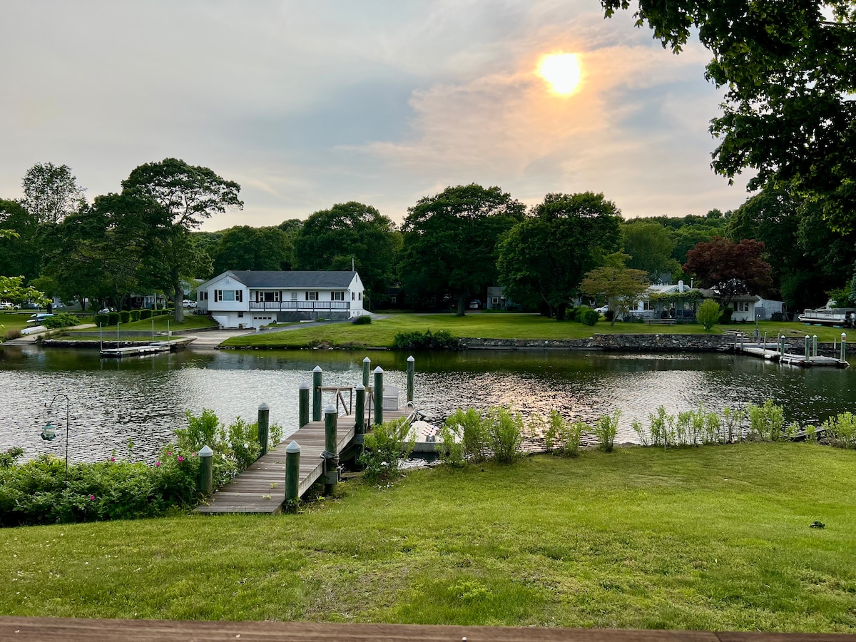 A serene riverfront view is presented, featuring a well-maintained dock stretching into calm waters. The sun sets softly behind a spacious single-level home, surrounded by lush greenery and mature trees. Various outdoor areas invite relaxation and enjoyment of the natural surroundings.