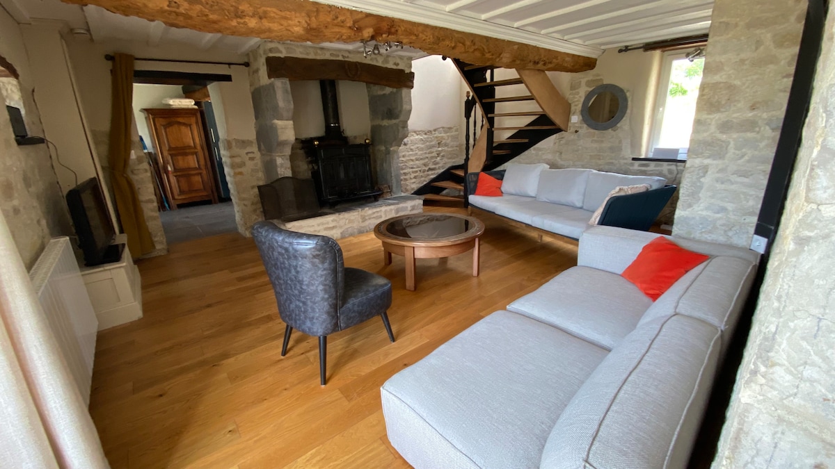 The living area features exposed wooden beams and a stone wall, creating a rustic charm. A light-colored sofa with red accent cushions and a modern armchair surround a circular wooden coffee table, with a fireplace positioned prominently in the corner.