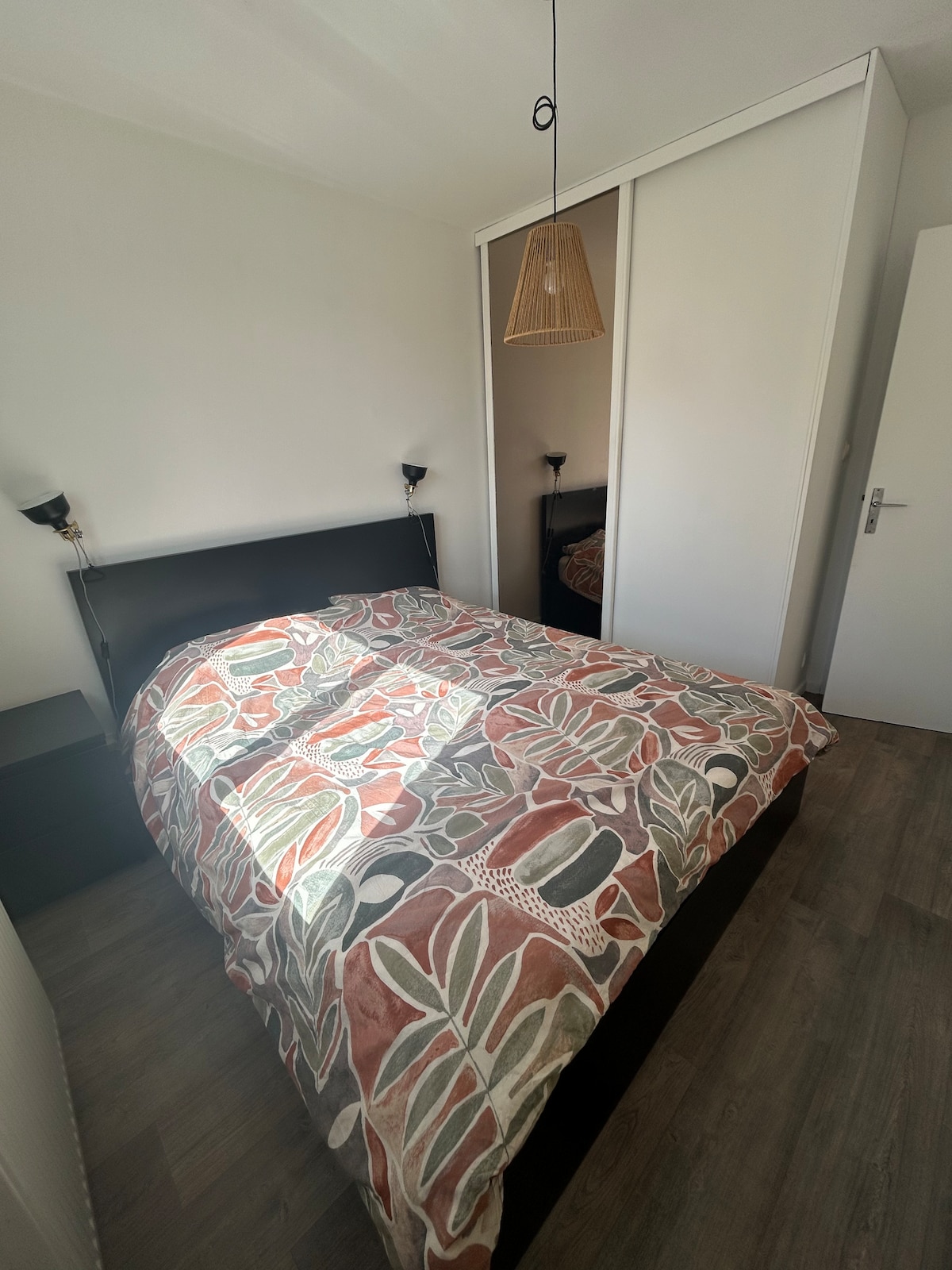A cozy bedroom is featured with a spacious bed adorned with a patterned duvet. A pendant light hangs above, illuminating the space. Two bedside lamps provide additional light, and a fitted wardrobe is visible to one side, enhancing the room's functional layout.