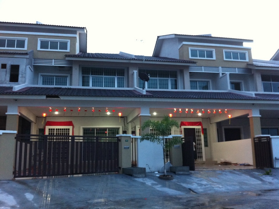 The exterior of a multi-story townhouse displays a modern design, featuring a warm color palette and a tiled roof. A welcoming entrance is framed by decorative lighting, and a gate leads to a landscaped area with a small garden. The setting is well-lit, suggesting a cheerful atmosphere.
