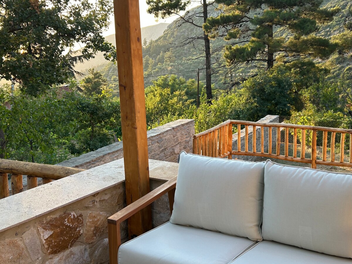 A cozy seating area is positioned under a wooden pergola, showcasing a comfortable sofa with soft cushions. Surrounding greenery and hills are visible in the background, creating a serene atmosphere. Natural light filters through the trees, enhancing the tranquil setting.