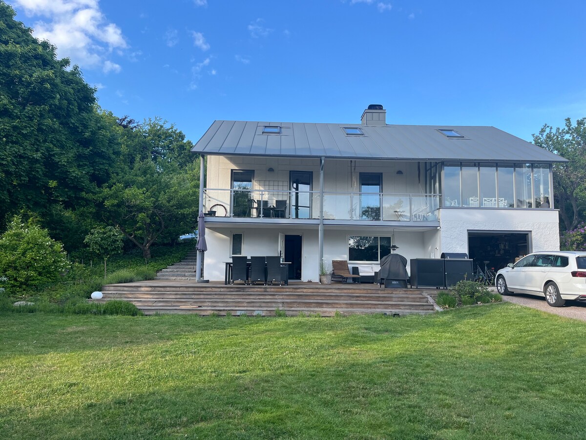 A modern house with a light-colored exterior is set against a clear blue sky. A spacious deck is seen in front, featuring outdoor seating arrangements. Lush greenery surrounds the property, enhancing its natural setting. Large windows provide ample natural light to the interior.