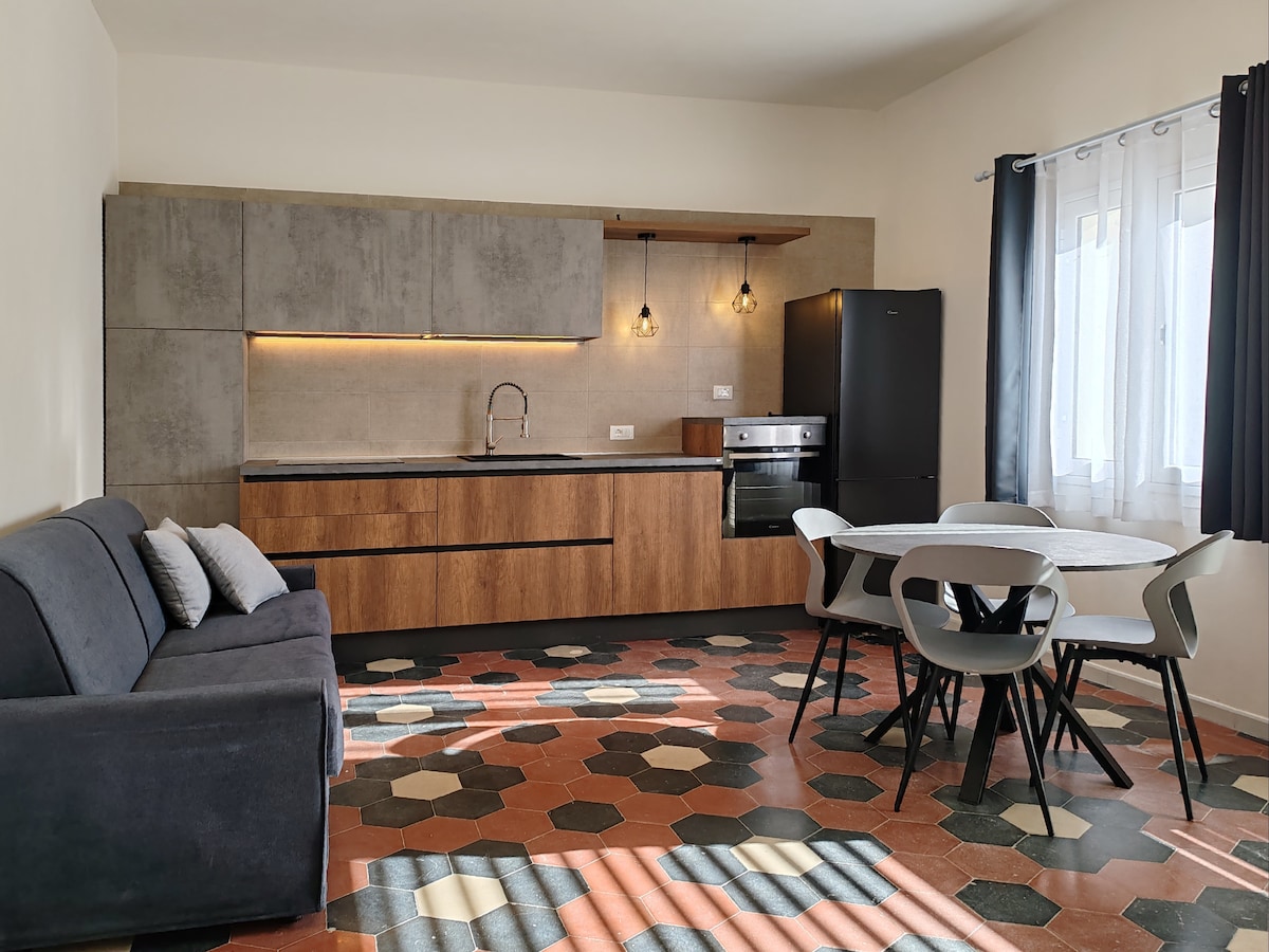 An indoor living area features a modern kitchen with sleek cabinetry and a dark refrigerator. A compact dining table with four chairs is positioned on a patterned tile floor, while a cozy sofa with decorative pillows is situated nearby. Natural light enters through two windows with elegant curtains.