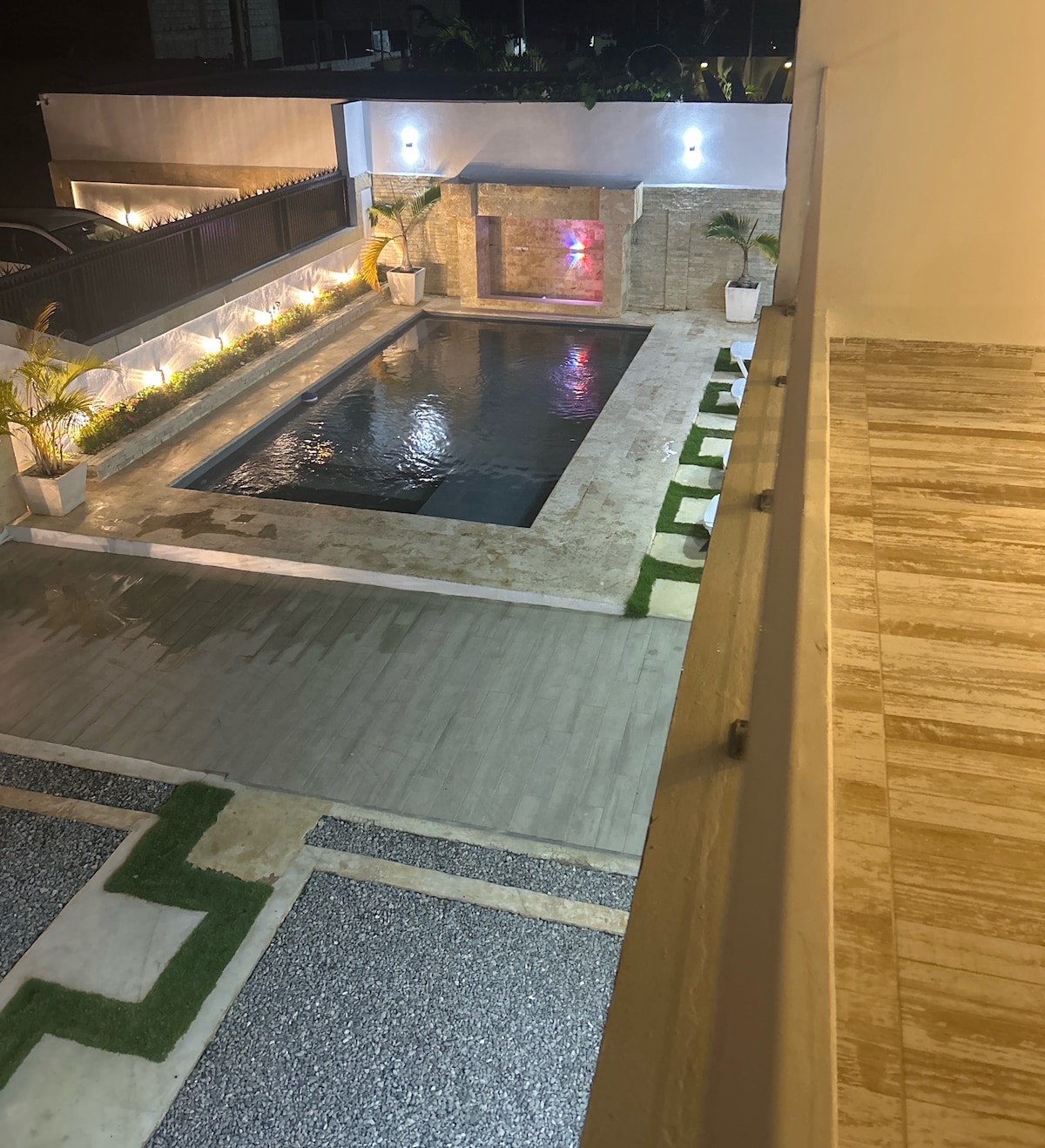 A spacious outdoor area features a pool surrounded by a well-maintained patio. Lounge chairs line the edge, with decorative plants adding a touch of greenery. Soft lighting enhances the tranquil atmosphere during the evening, creating a welcoming setting for relaxation.