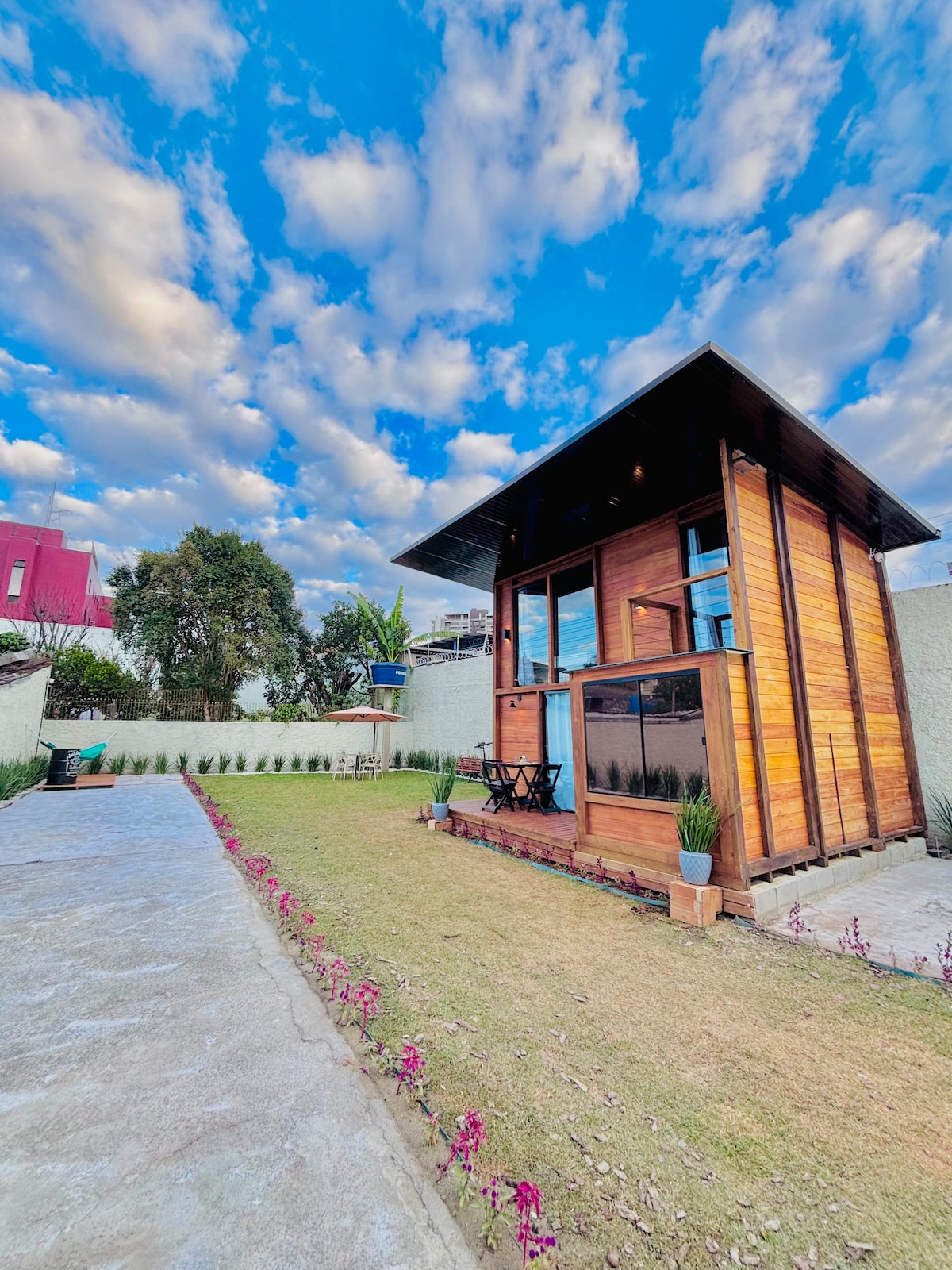 A charming wooden structure is set against a dynamic sky filled with fluffy clouds. The small building features large windows and a cozy exterior. A well-maintained garden with a grassy area and flower borders runs alongside a stone pathway leading to the entrance.
