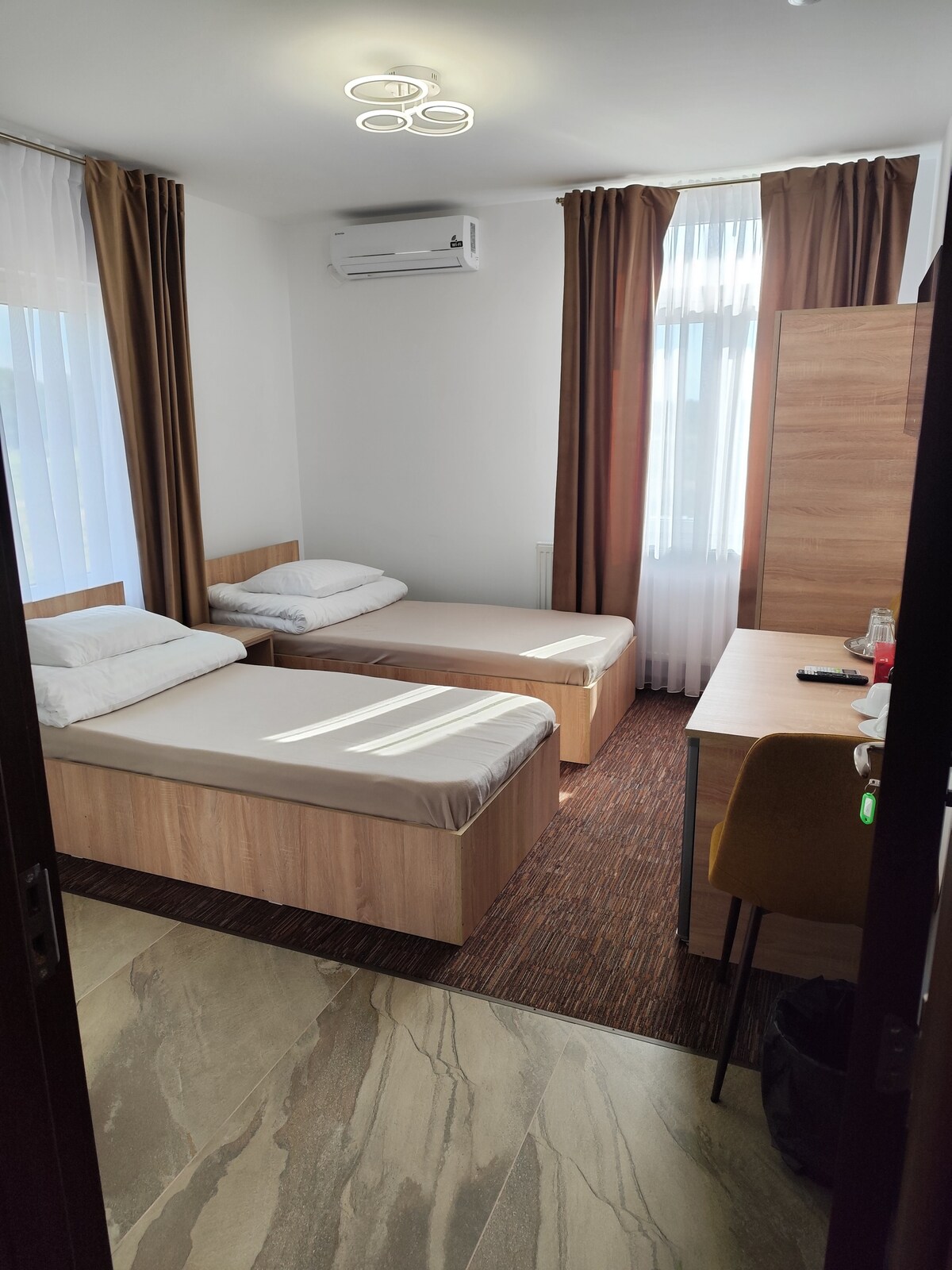 A well-lit guest room features two twin beds with neutral bedding, positioned against a light-colored wall. Natural light filters through sheer curtains, and a desk with a chair is placed near the wall. Air conditioning is installed on the wall for added comfort.
